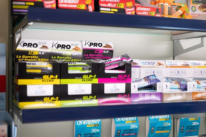 Una persona in camice bianco, con le braccia incrociate, tiene in mano uno stetoscopio, sullo sfondo sfocato di una farmacia.