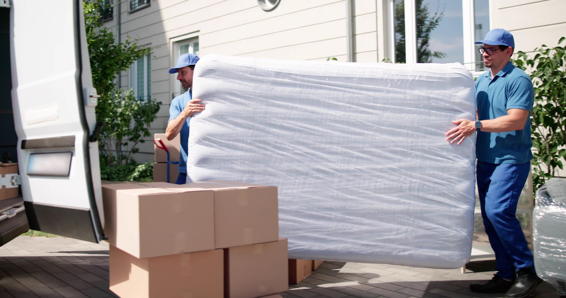 Two movers in blue unloading a wrapped mattress from a van, boxes on the ground.