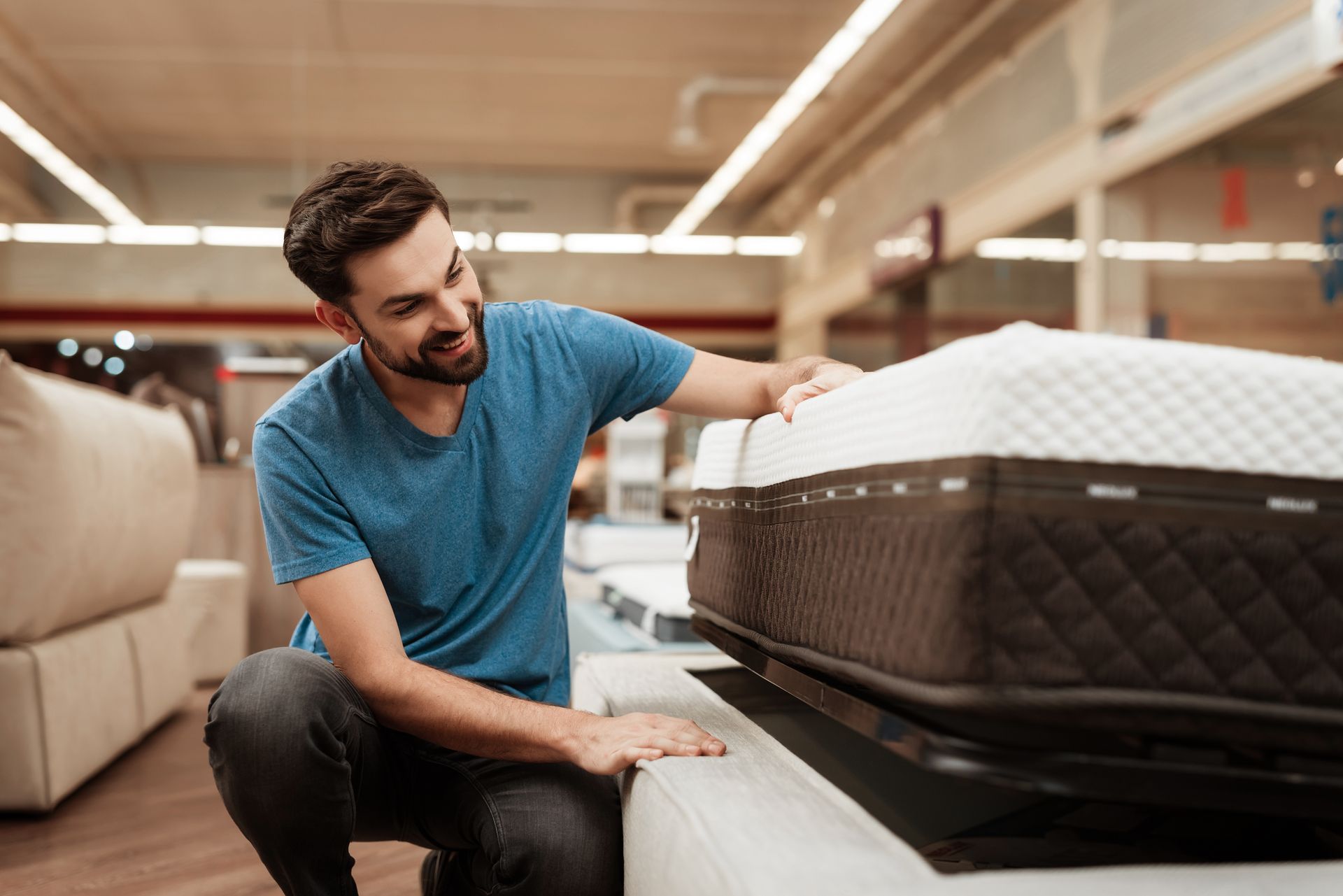Man kneels in a store, examining a mattress. He smiles, touching the mattress.