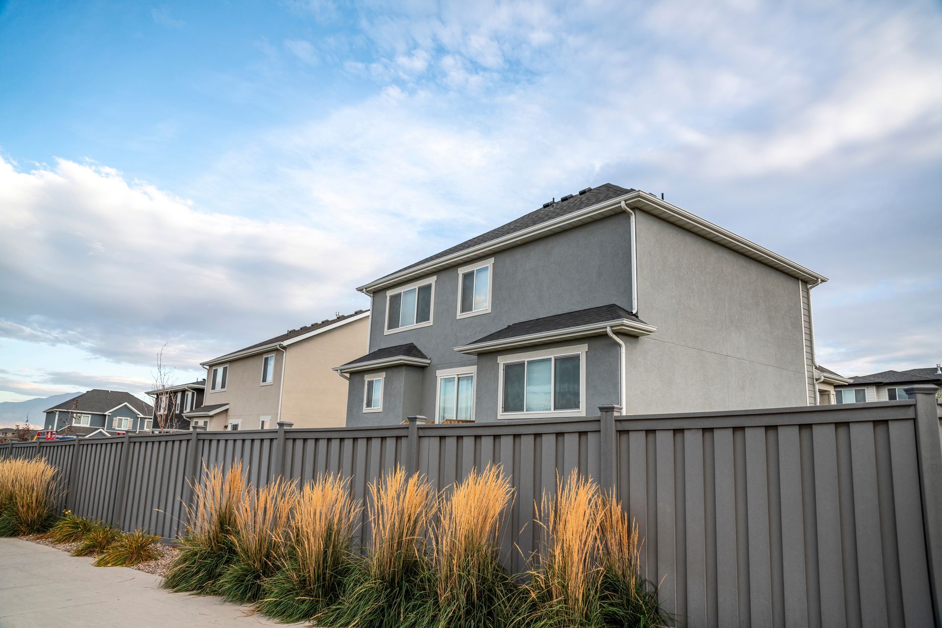 Two-story houses with gray siding behind a gray wooden fence and tall grasses under a cloudy blue sky.