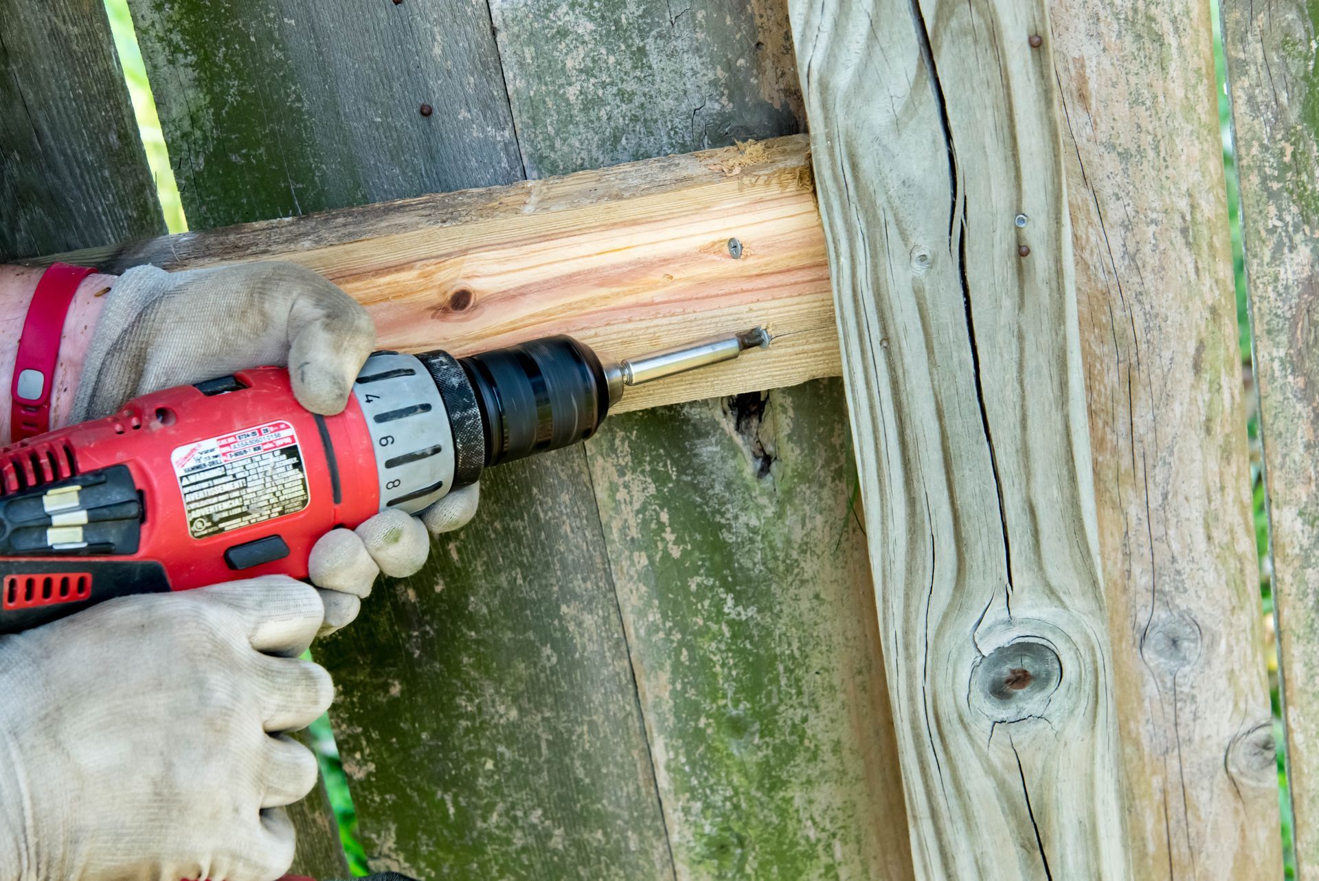 Person wearing gloves using a red drill to screw into a wooden fence.