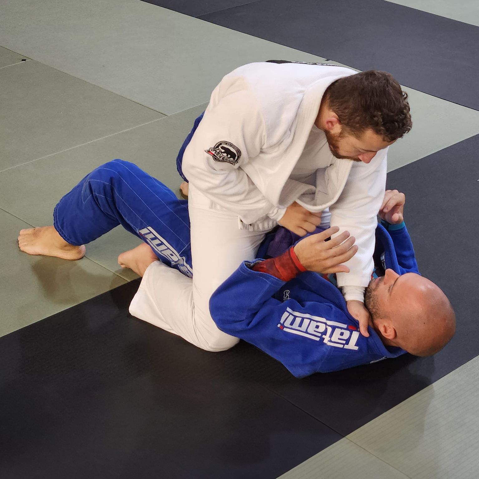 Two men are practicing jiu jitsu and one of them is wearing a blue uniform that says tatami