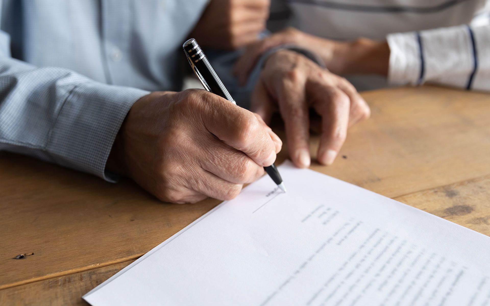 Person signing a document with a pen, another person's arm in the background; both are at a table.