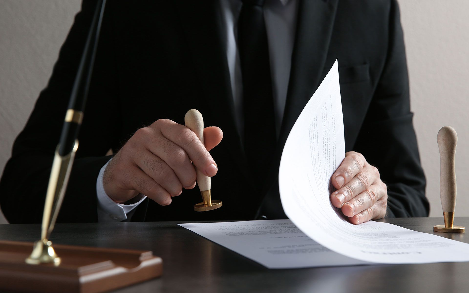 Person in suit stamps a document with a wooden stamp. A pen and another stamp are on the desk.