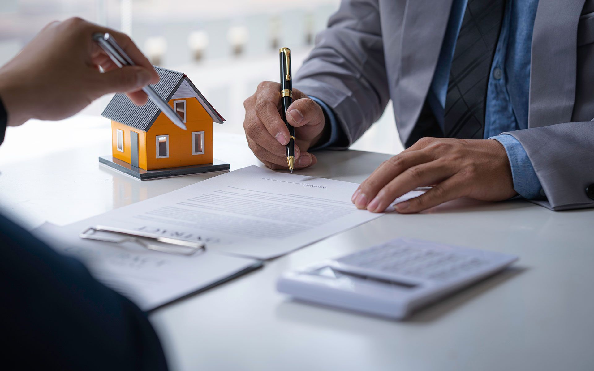 Two people signing a document at a desk, with a model house and calculator present; suggesting a real estate transaction.