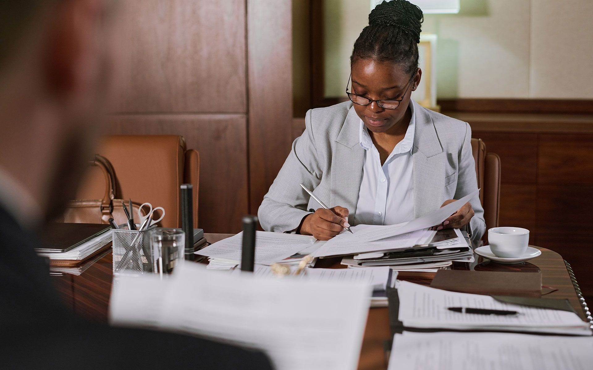 A Black woman in a light grey blazer sits at a table, reviewing documents, in a wood-paneled office.