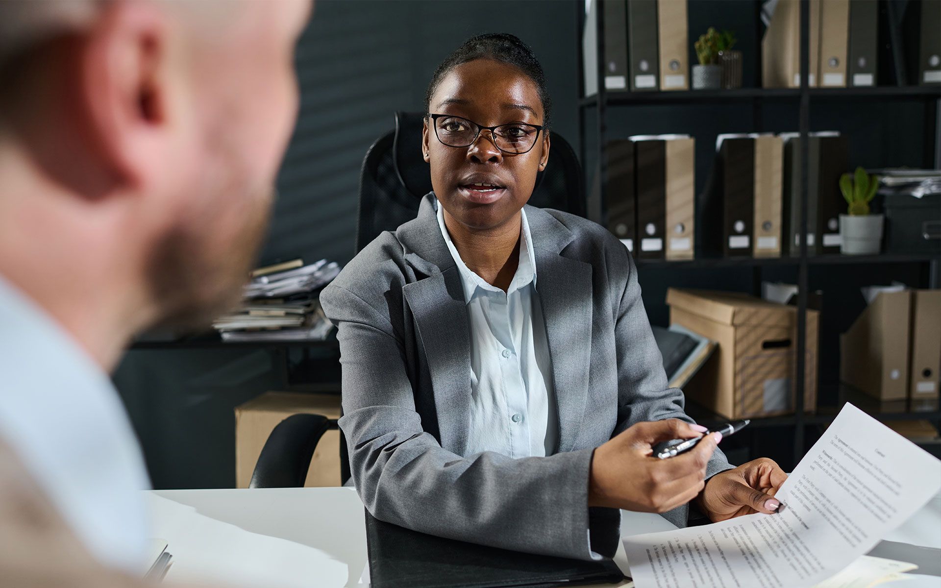 A Black woman in a grey suit reviews paperwork with a man at a desk in an office.