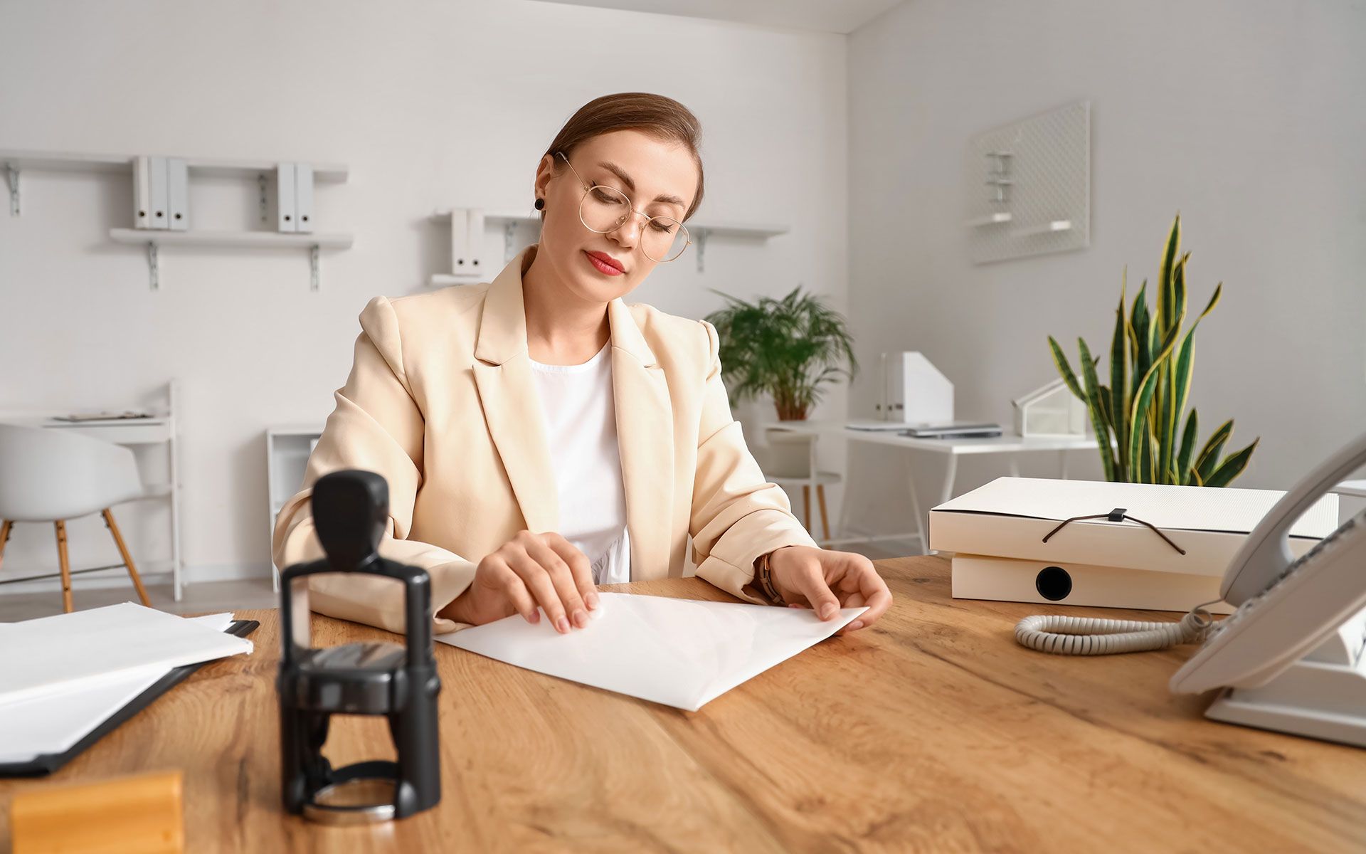 Woman in a beige blazer at a wooden desk, examining a document with a stamp nearby in an office setting.