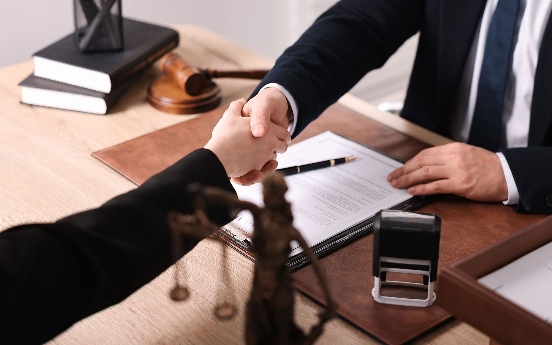 Two people in suits shaking hands over a document, with a gavel, books, and a statue of Lady Justice visible on a desk.