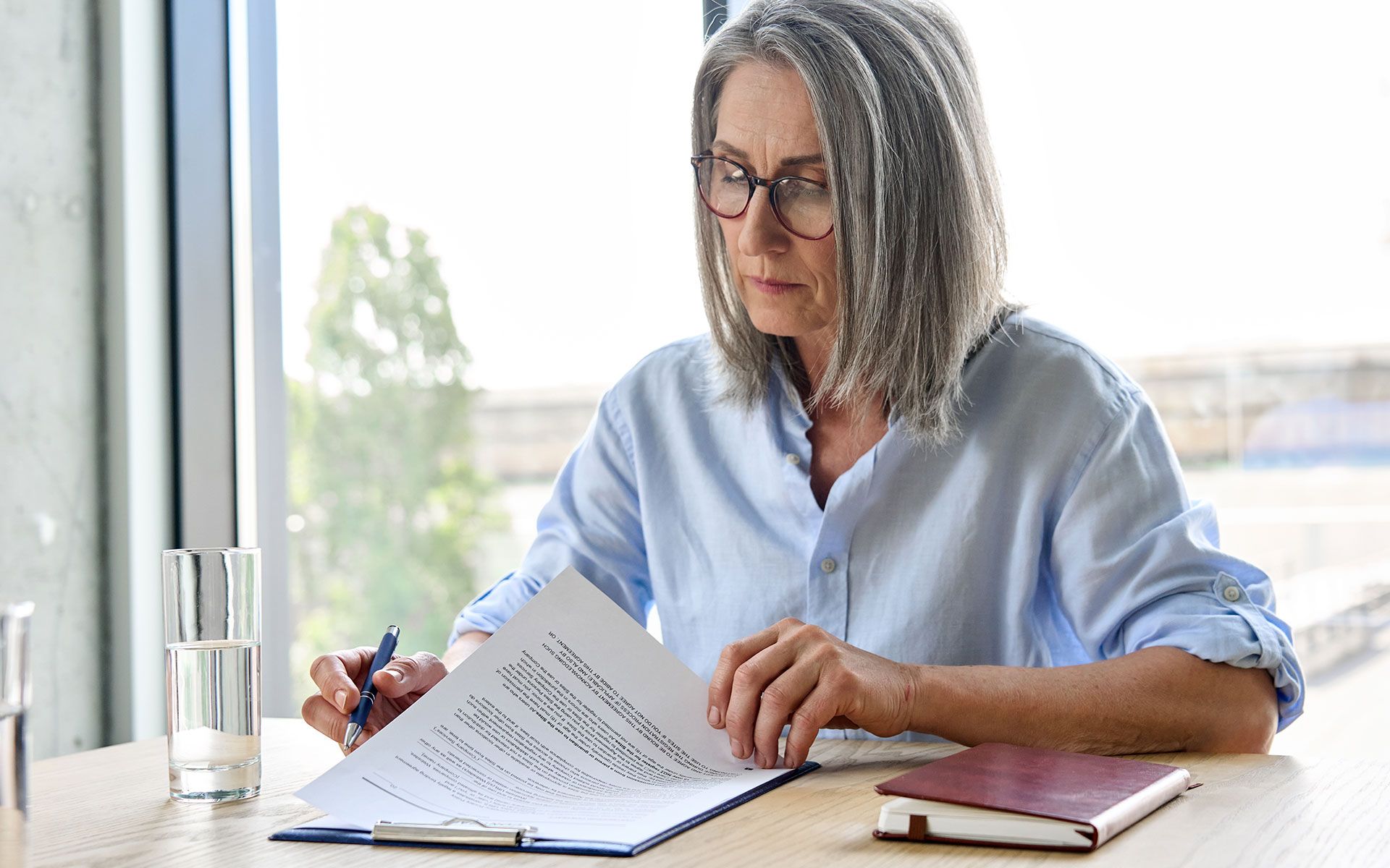 Woman with gray hair and glasses reviewing documents at a table, holding a pen.