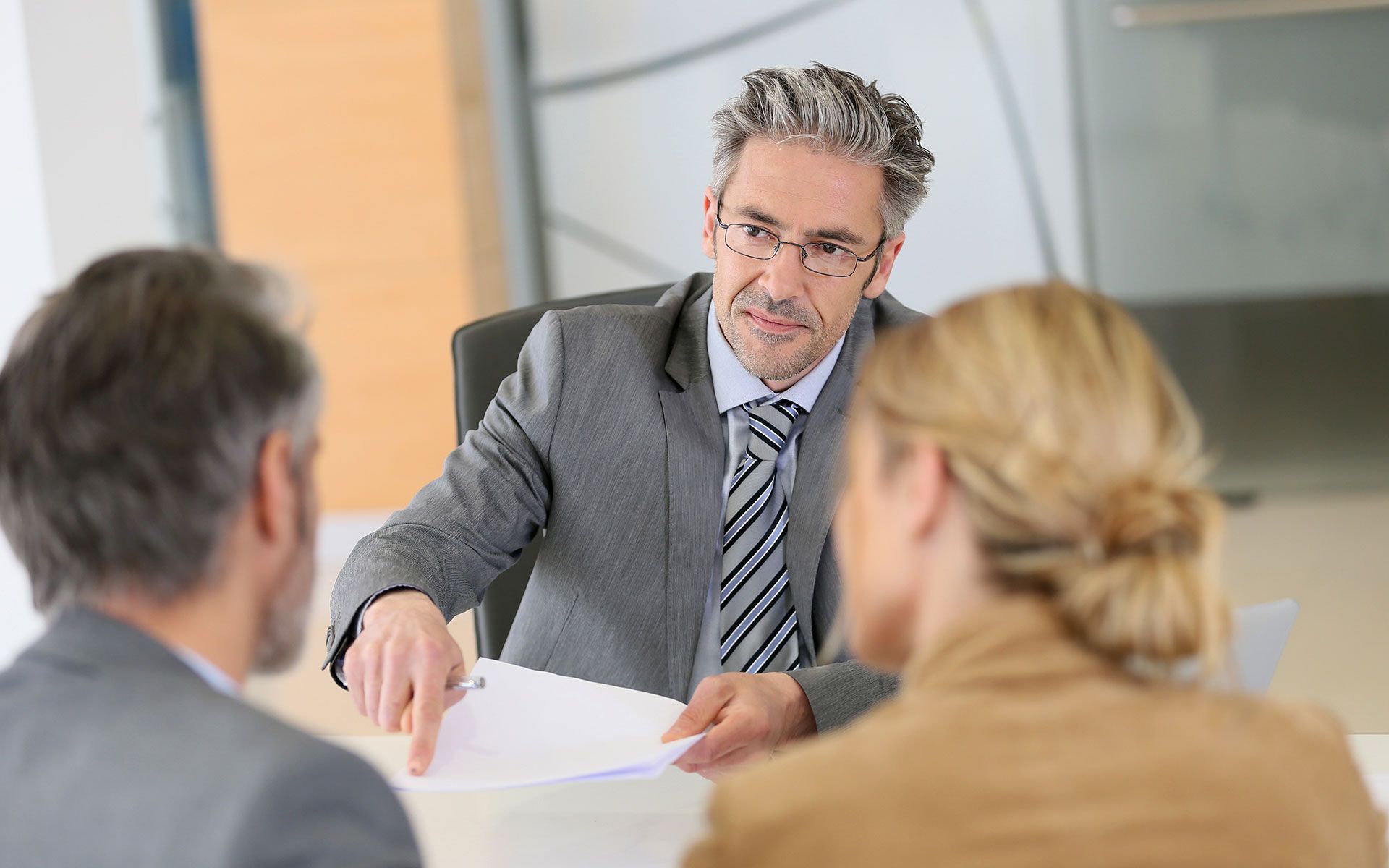 A man in a suit points to documents as he advises a couple, all seated at a desk in an office.