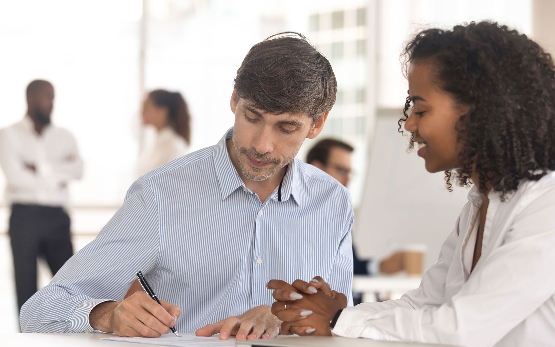 A man signing a document while a woman watches and smiles in a bright office setting.
