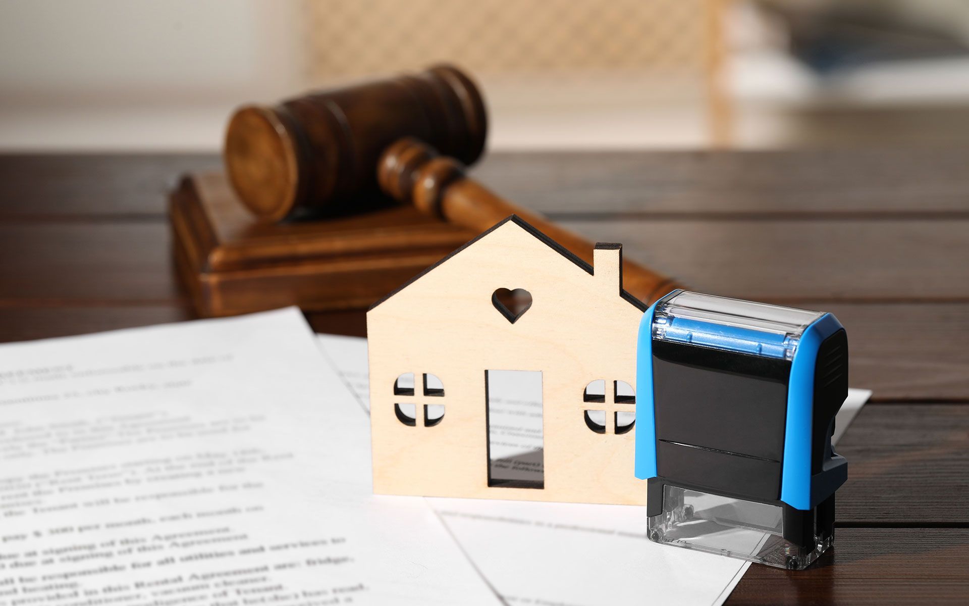 A wooden house cutout, documents, a gavel, and a stamp on a wooden table, symbolizing real estate or legal proceedings.