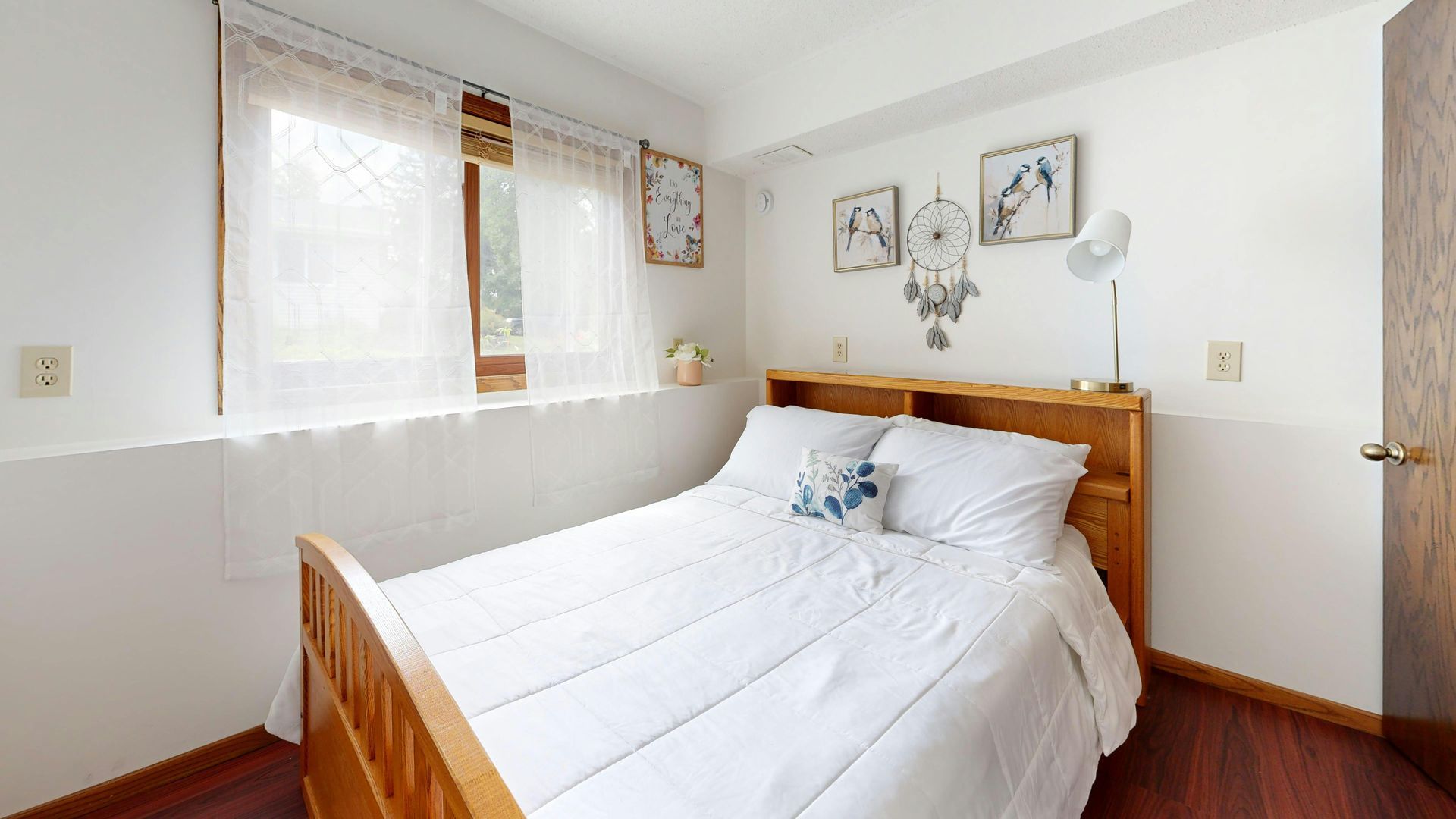 Bedroom with a white bed, wooden headboard, small window with curtains, and wall art.