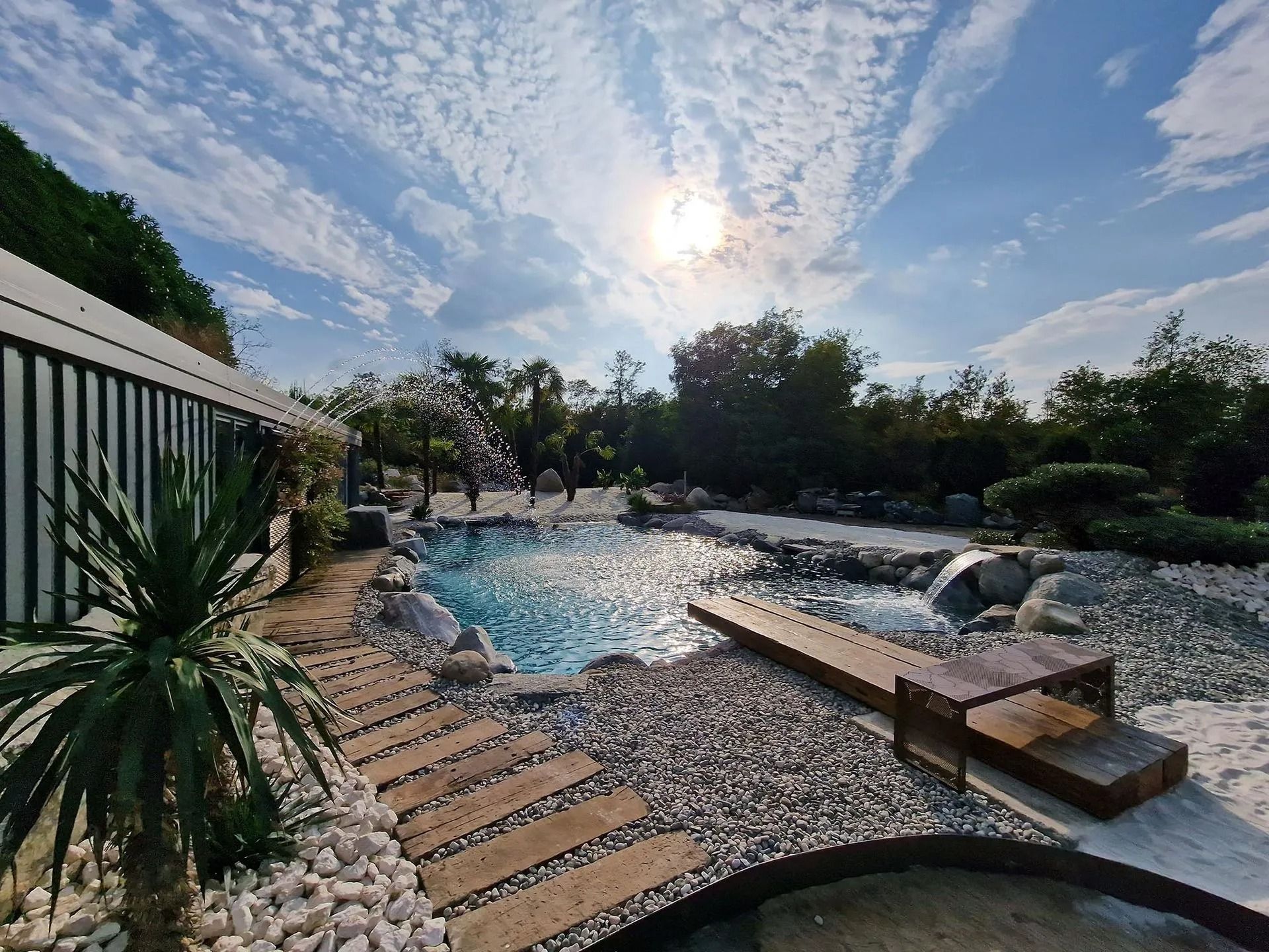 Piscina con giochi d'acqua, ponte di legno, rocce e un rigoglioso giardino sotto un cielo soleggiato.