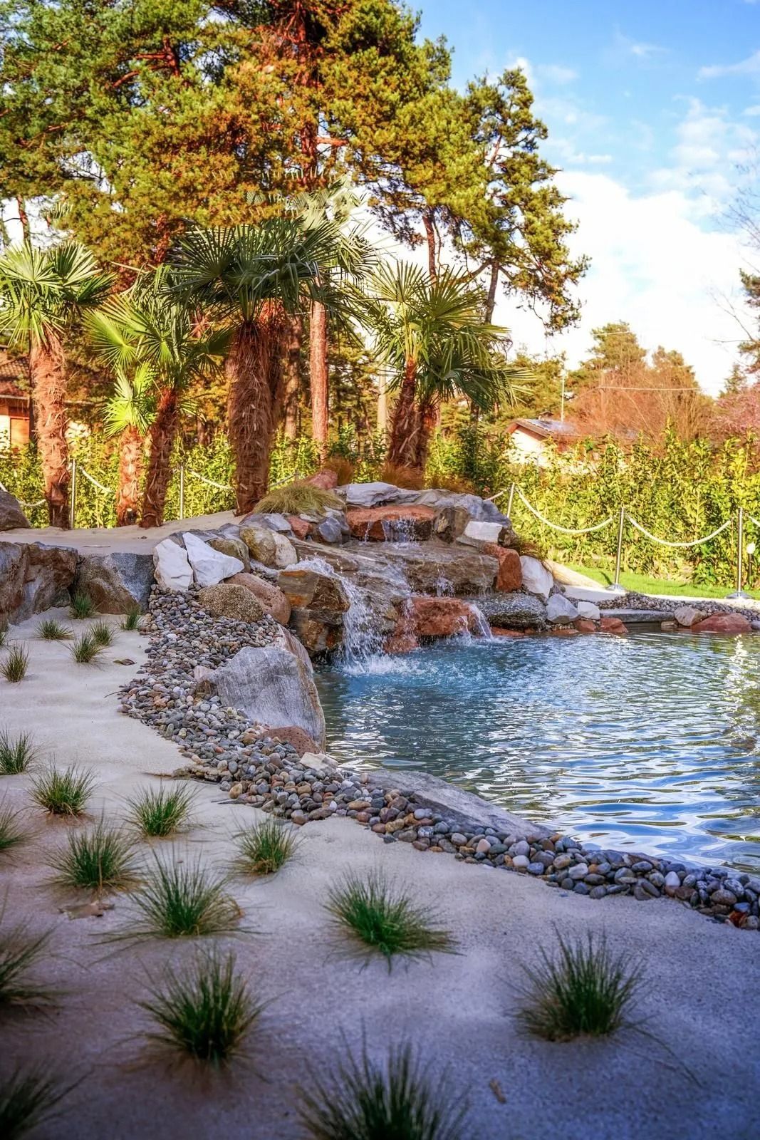 Un laghetto tranquillo con una piccola cascata, circondato da alberi, rocce e sabbia.