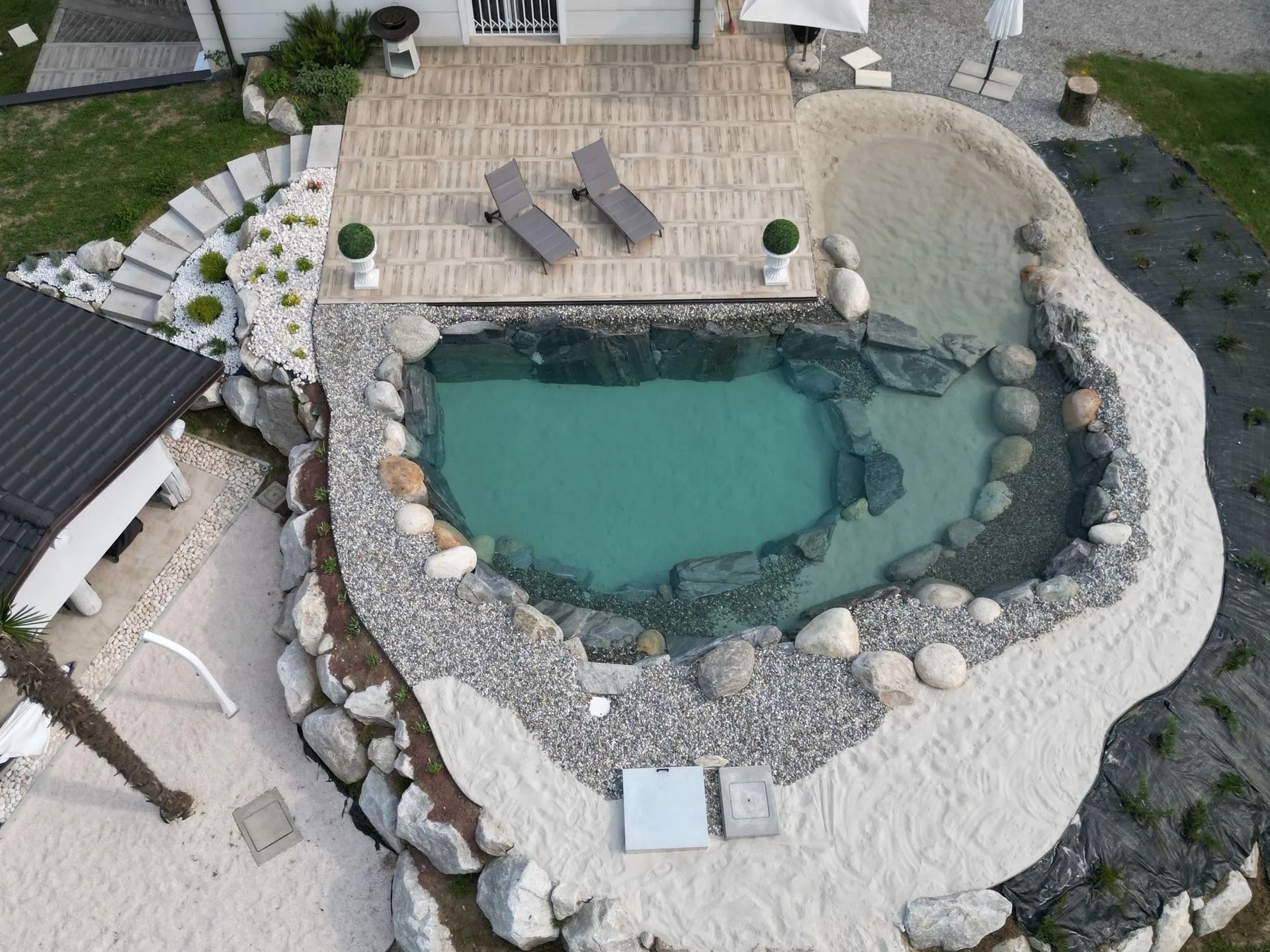 Vista aerea di una piscina dall'aspetto naturale con spiaggia sabbiosa ed elementi in pietra. Due sedie a sdraio su una terrazza in legno.