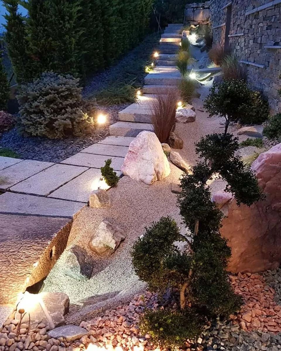 Un sentiero in pietra con illuminazione integrata sale verso un giardino paesaggistico, caratterizzato da rocce, arbusti e un bonsai.