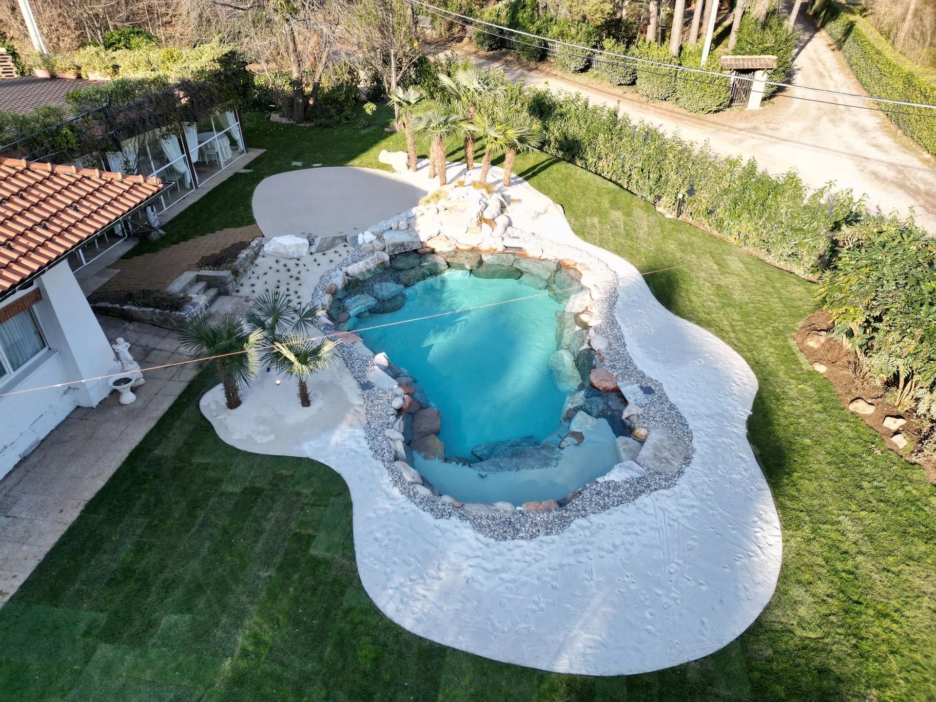 Vista aerea di una piscina personalizzata a forma libera, circondata da rocce, sabbia bianca e un rigoglioso prato verde.