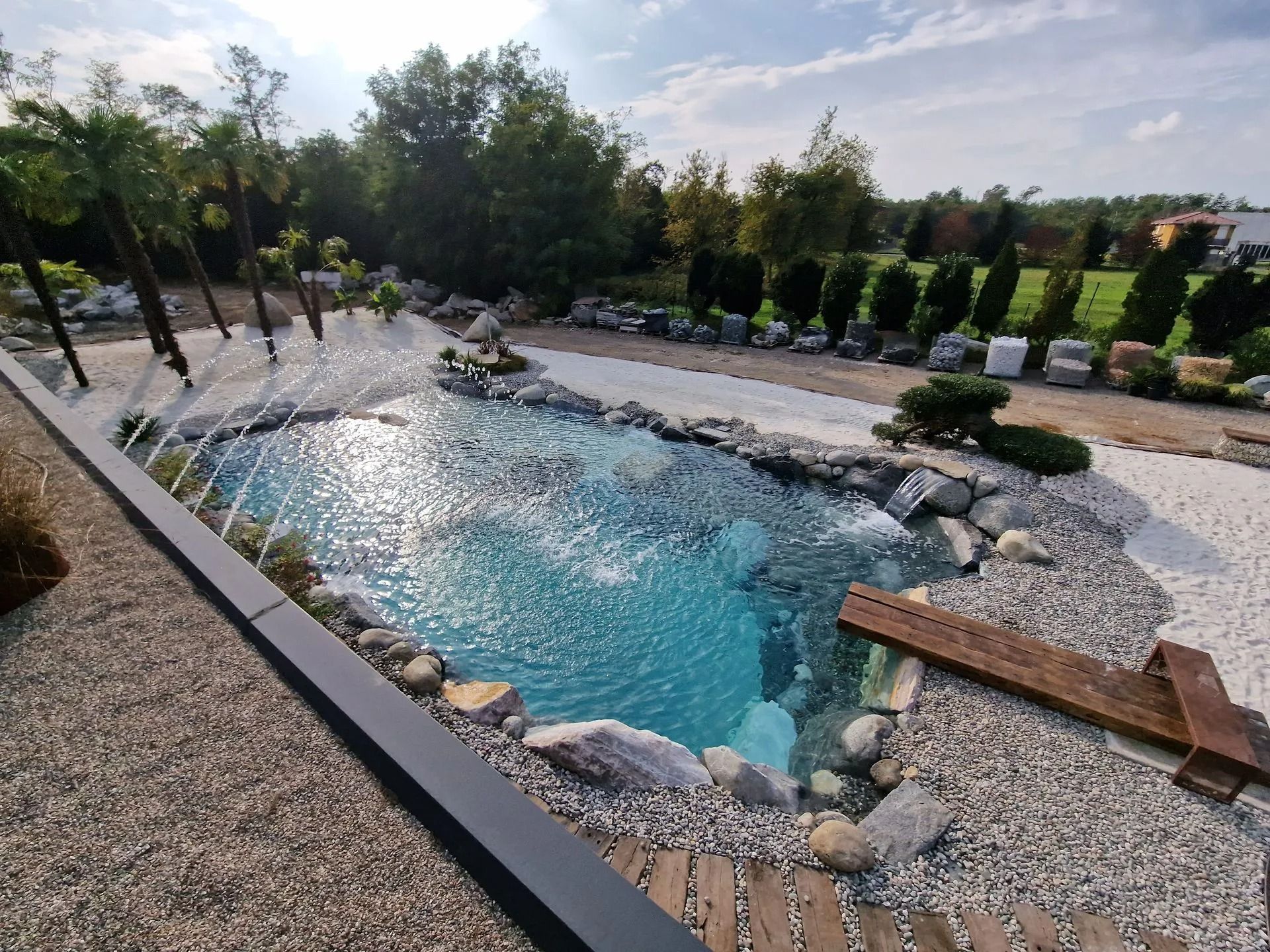 Uno stagno balneabile dalle acque azzurre con rocce, un trampolino e ciottoli bianchi circostanti, sotto un cielo parzialmente nuvoloso.