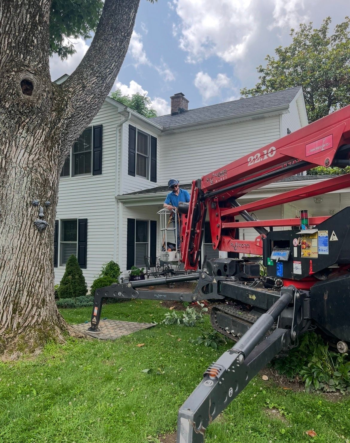 Man in a lift trims tree branches near a white house with black shutters on a sunny day.