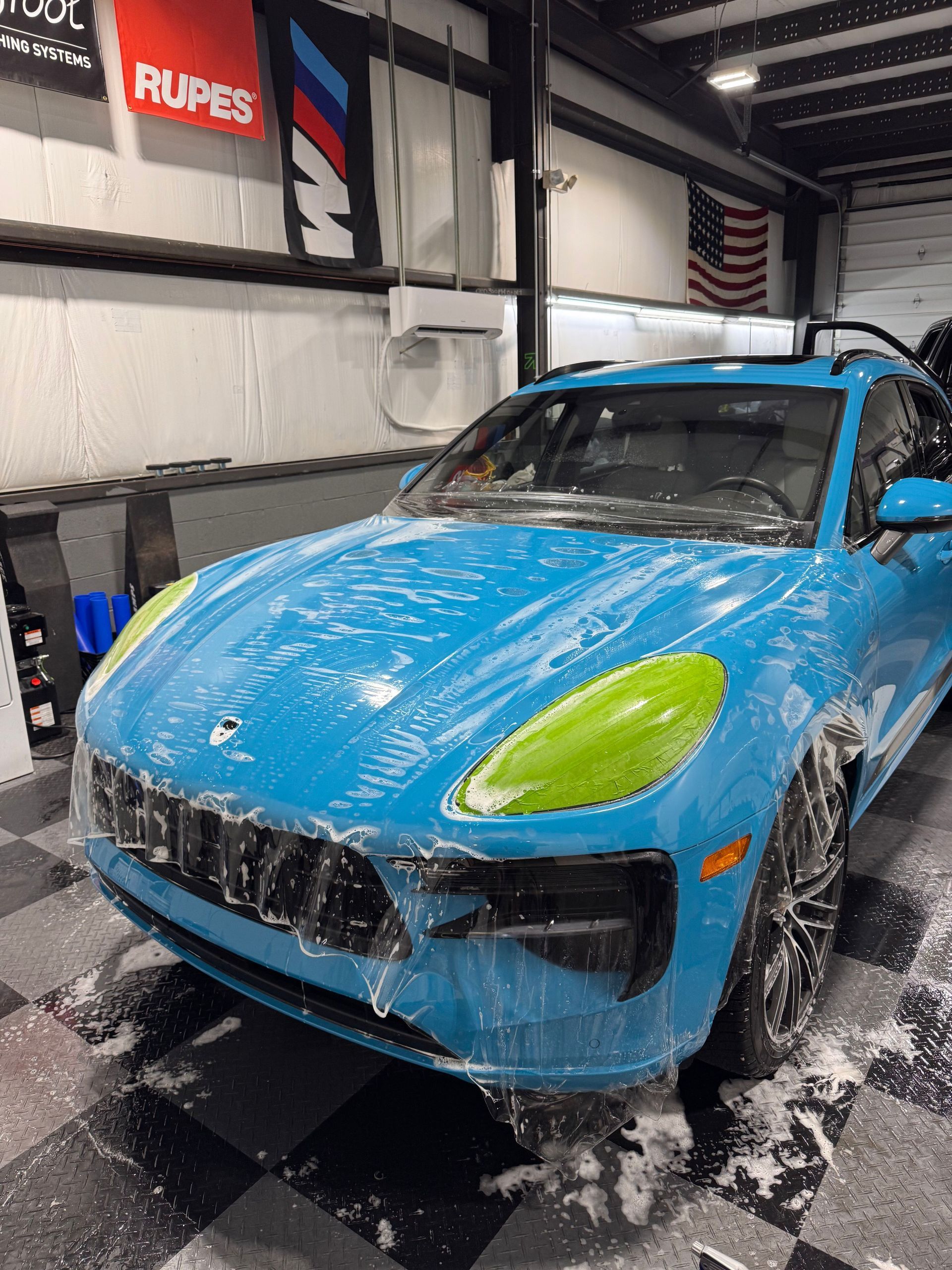 A bright blue Porsche Macan covered in soap suds sits inside a garage, with green tape applied to the headlights.