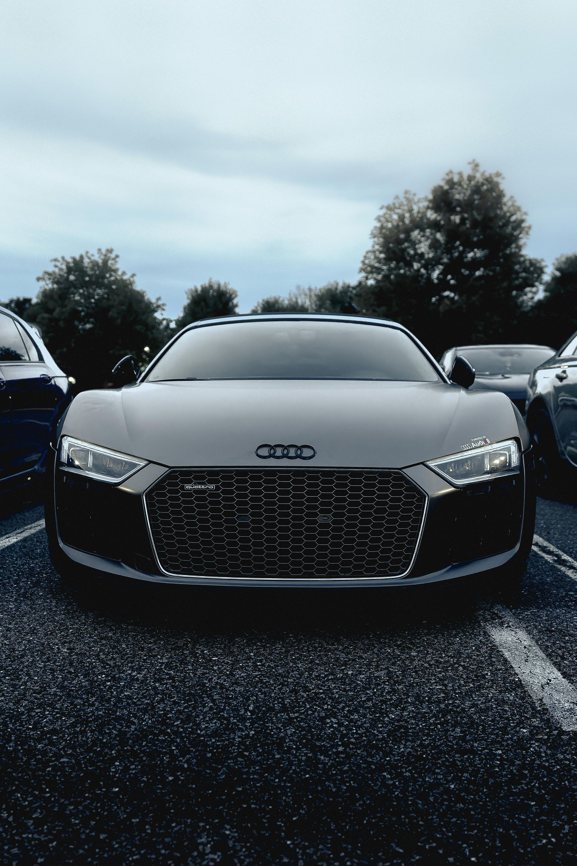 A dark gray Audi R8 sports car parked in an outdoor lot during an overcast day.