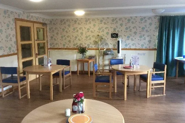 A furnished room with tables, chairs, and floral wallpaper. Sunlight streams through a window.