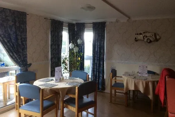 Corner dining room with tables, blue chairs, patterned wallpaper, and curtains; decorative car art on wall.