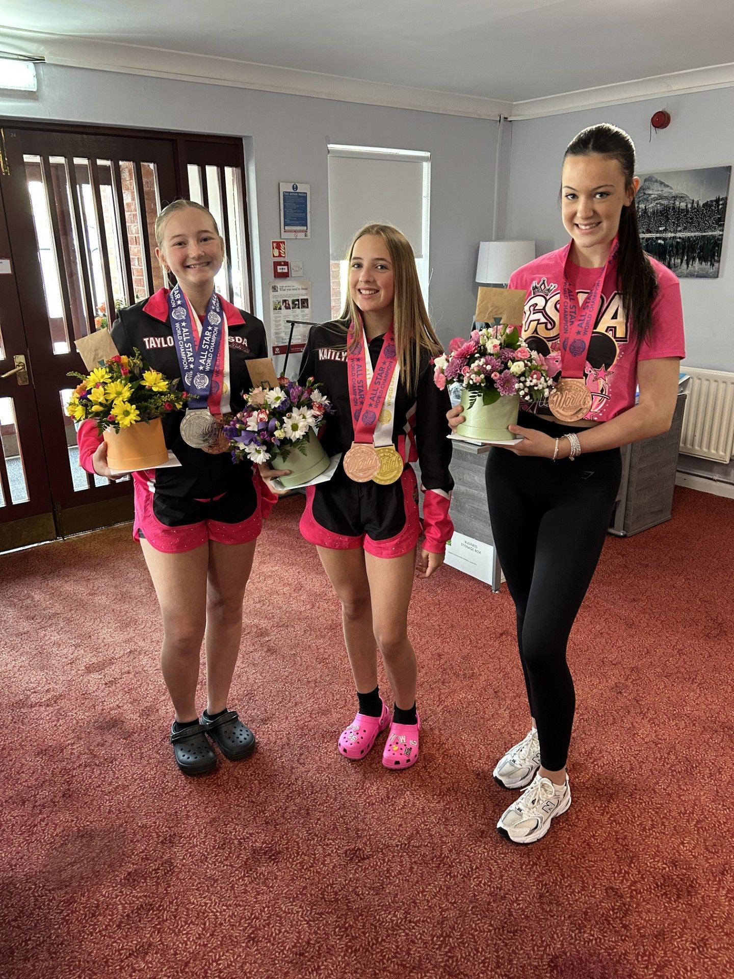 Three young women are holding flowers and medals in a living room.