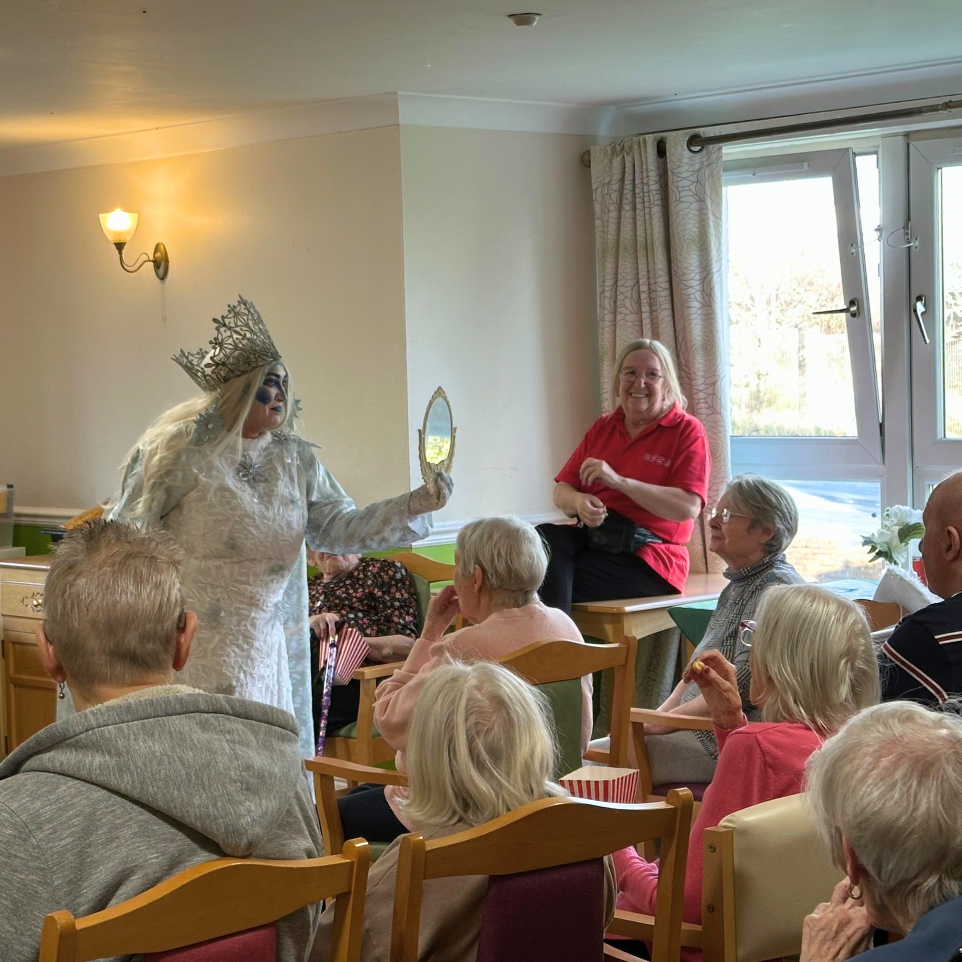 Person in costume holds a mirror while entertaining a group of seated people in a brightly lit room.