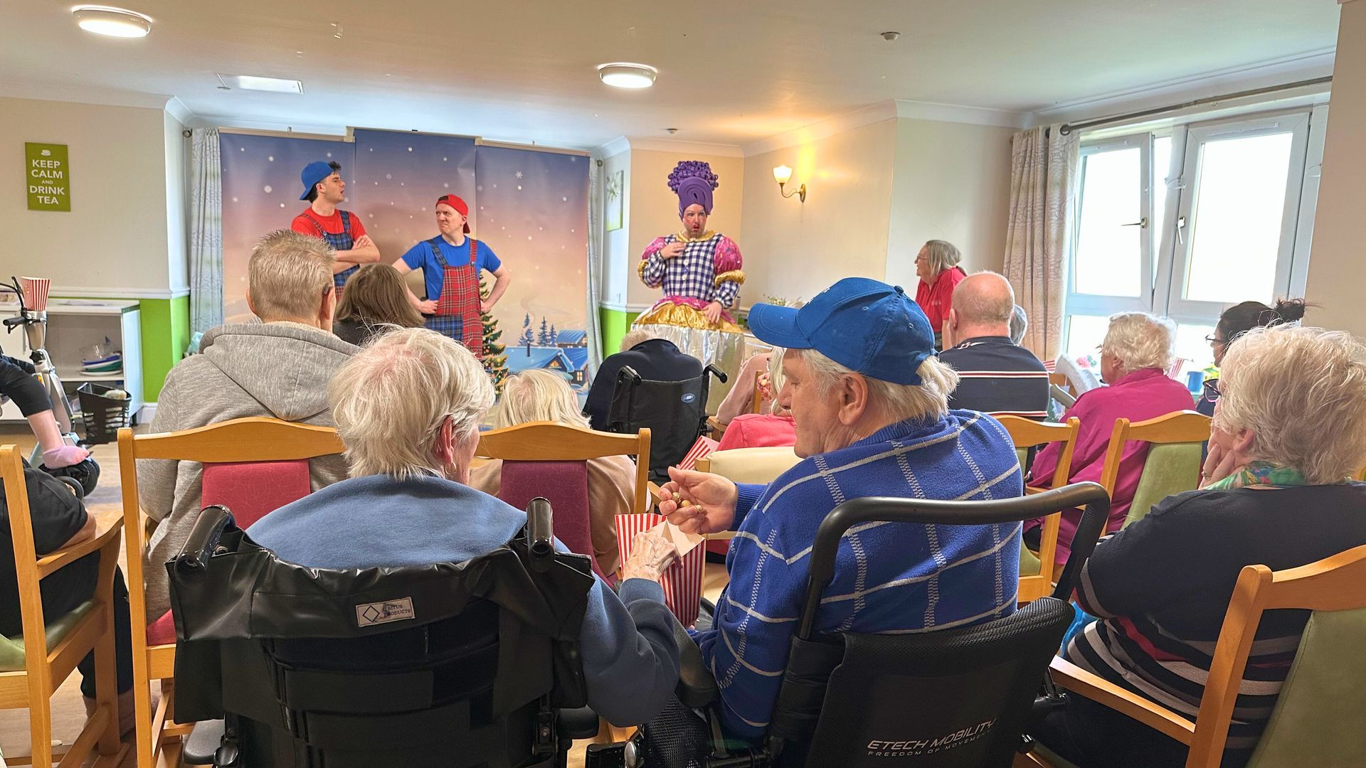 Audience watches performers on stage at an event. Stage has backdrop. People in wheelchairs and chairs.