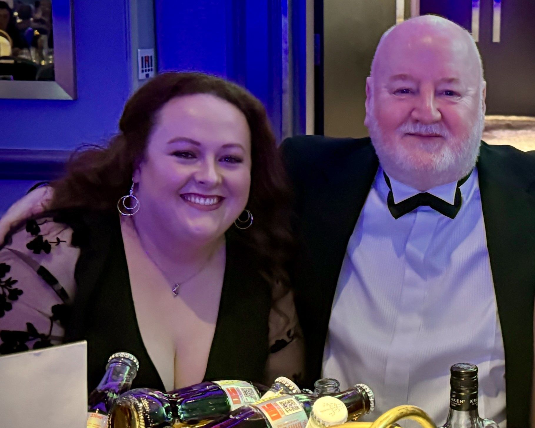 Woman and man smiling, seated at a table with drinks, in a room with blue lighting.