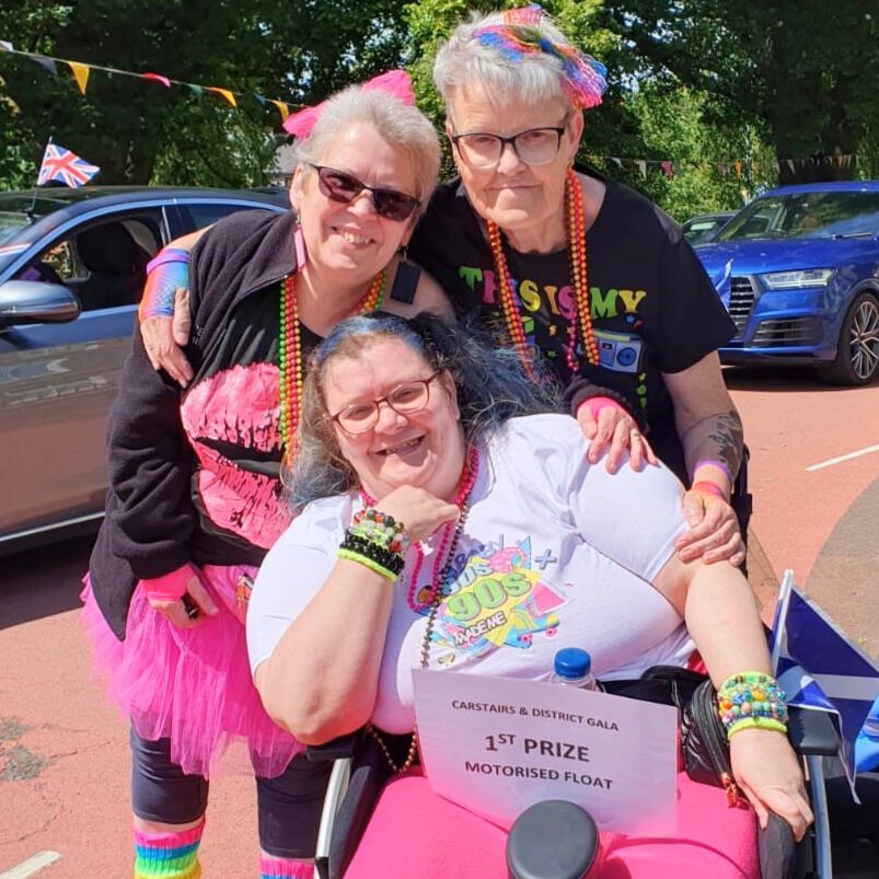 Three smiling people, two flanking one in a wheelchair, pose outdoors at an event. They wear brightly colored 80s-themed outfits, with a sign indicating a first-place prize.