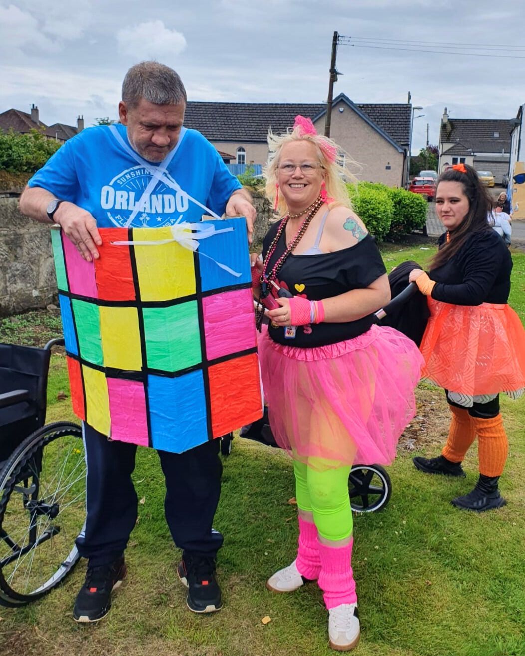 Man in a Rubik's Cube costume, woman in neon 80s attire, and another woman pushing a stroller. Outdoors, sunny.
