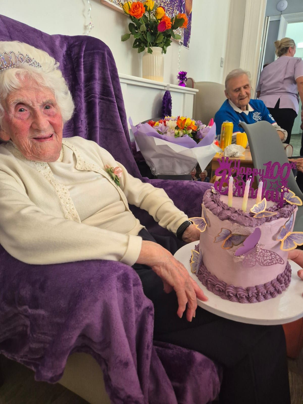 Elderly woman with tiara, holding cake, celebrating a birthday. Purple theme, flowers, and other people.