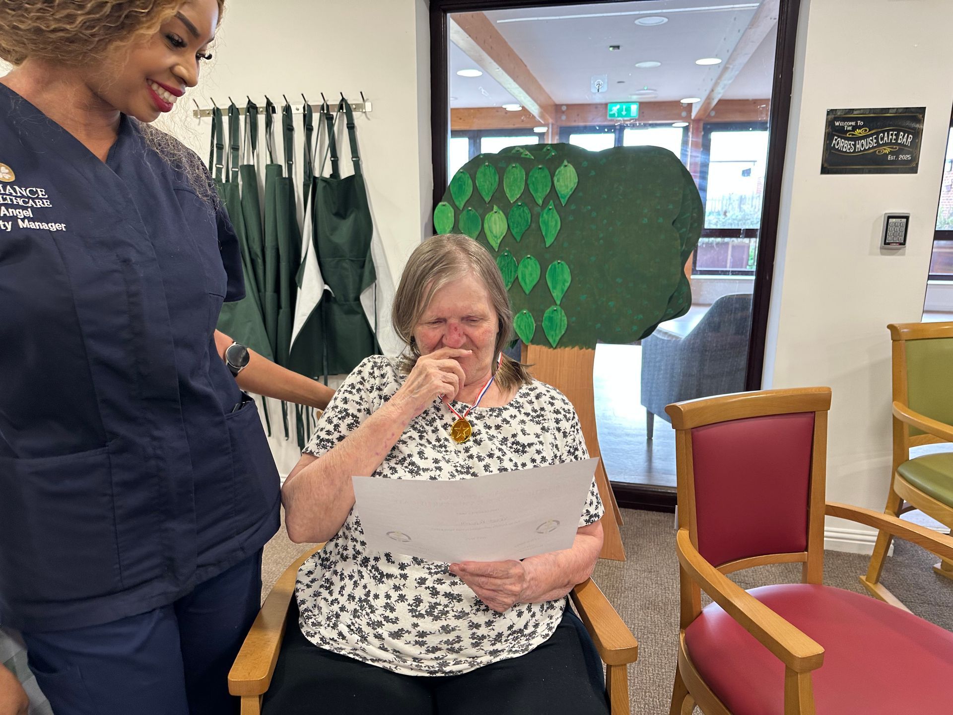 Woman in patterned top laughs while reading paper; a person in scrubs stands beside her.