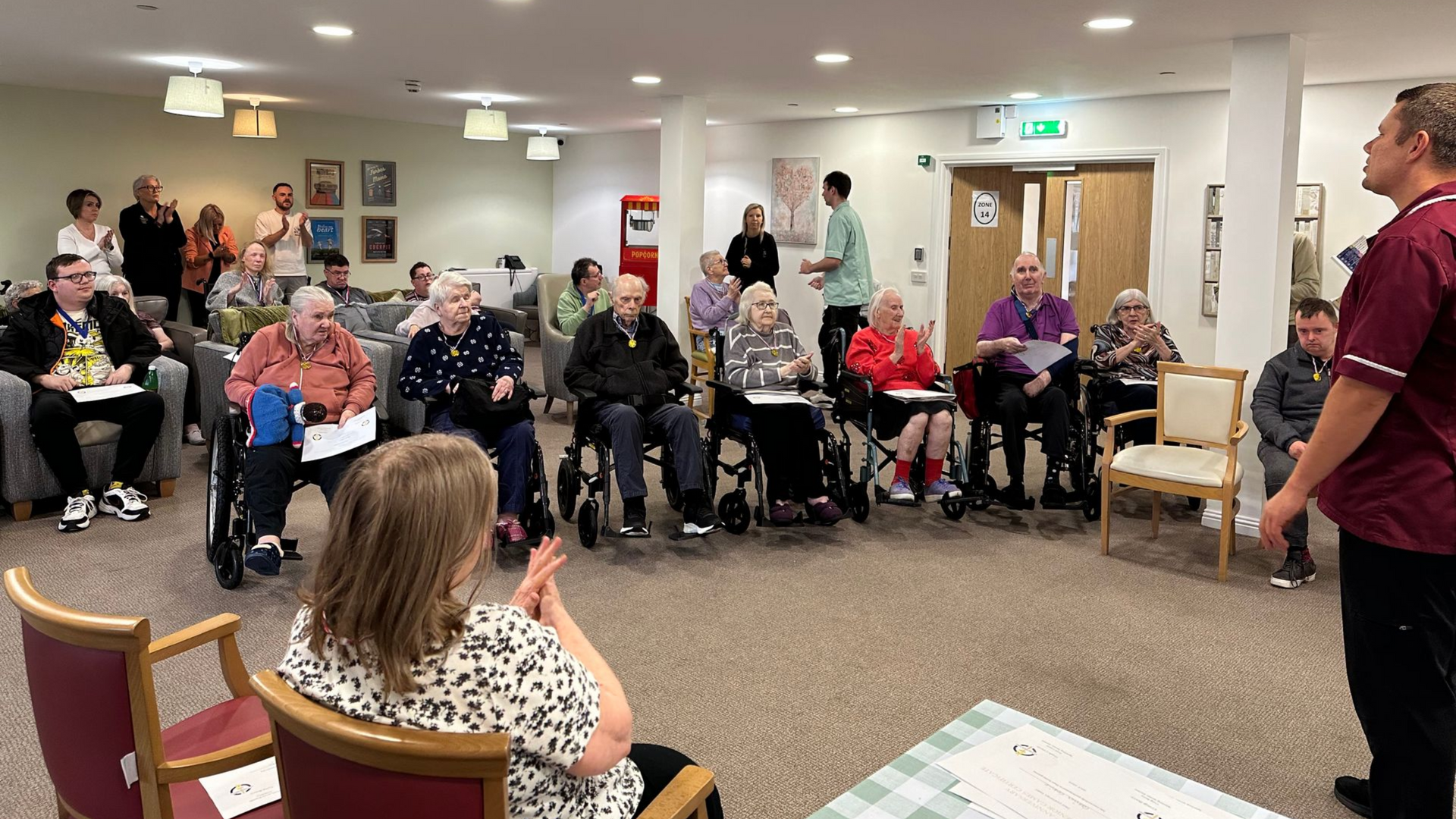 Group of people in a room, some in wheelchairs. A staff member speaks to the group. Another person claps.