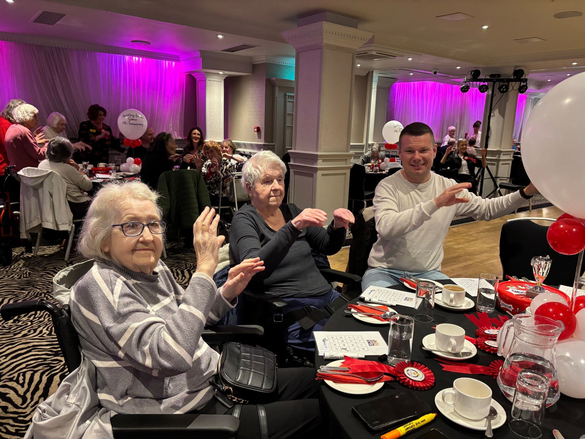 People at a table in a decorated room, smiling and clapping. Red and white balloons.