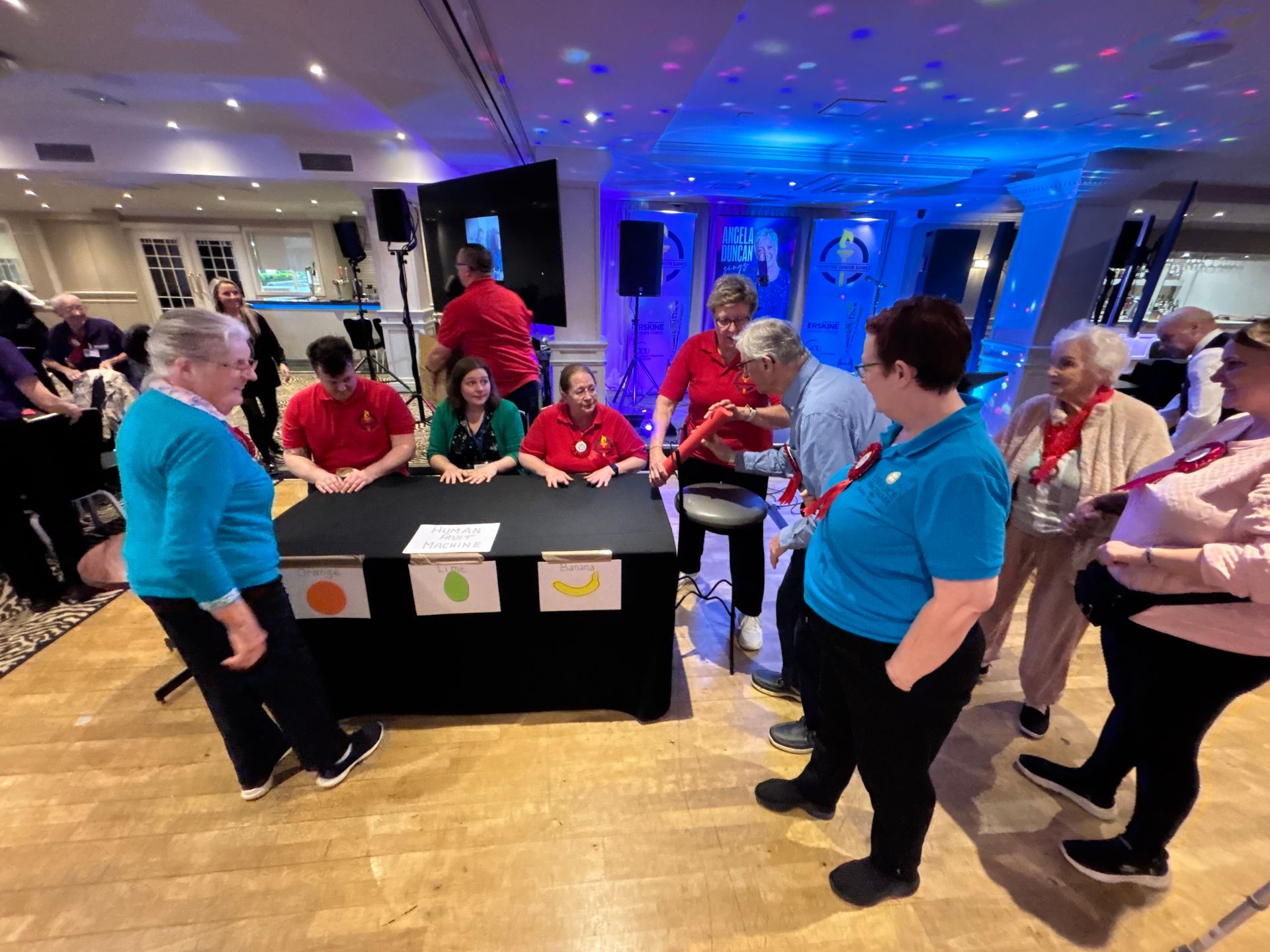 People at a party, some wearing red and teal shirts, around a table with fruit drawings.