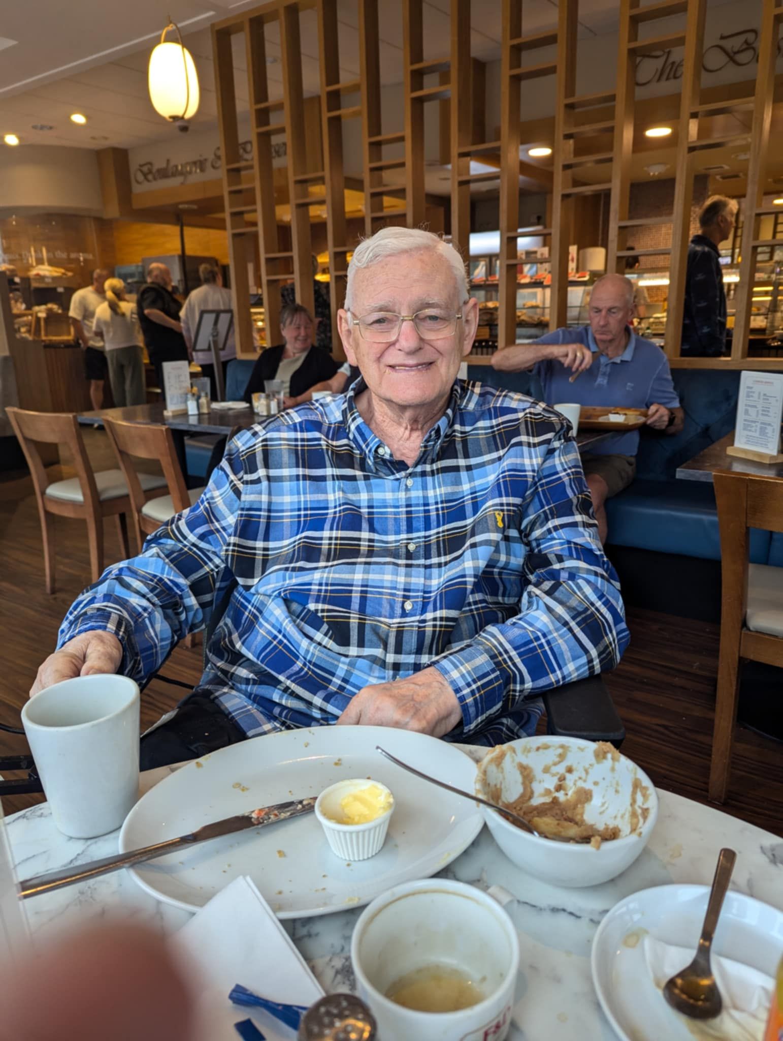 Elderly man smiles at a table with food and coffee. Restaurant setting, blue plaid shirt.