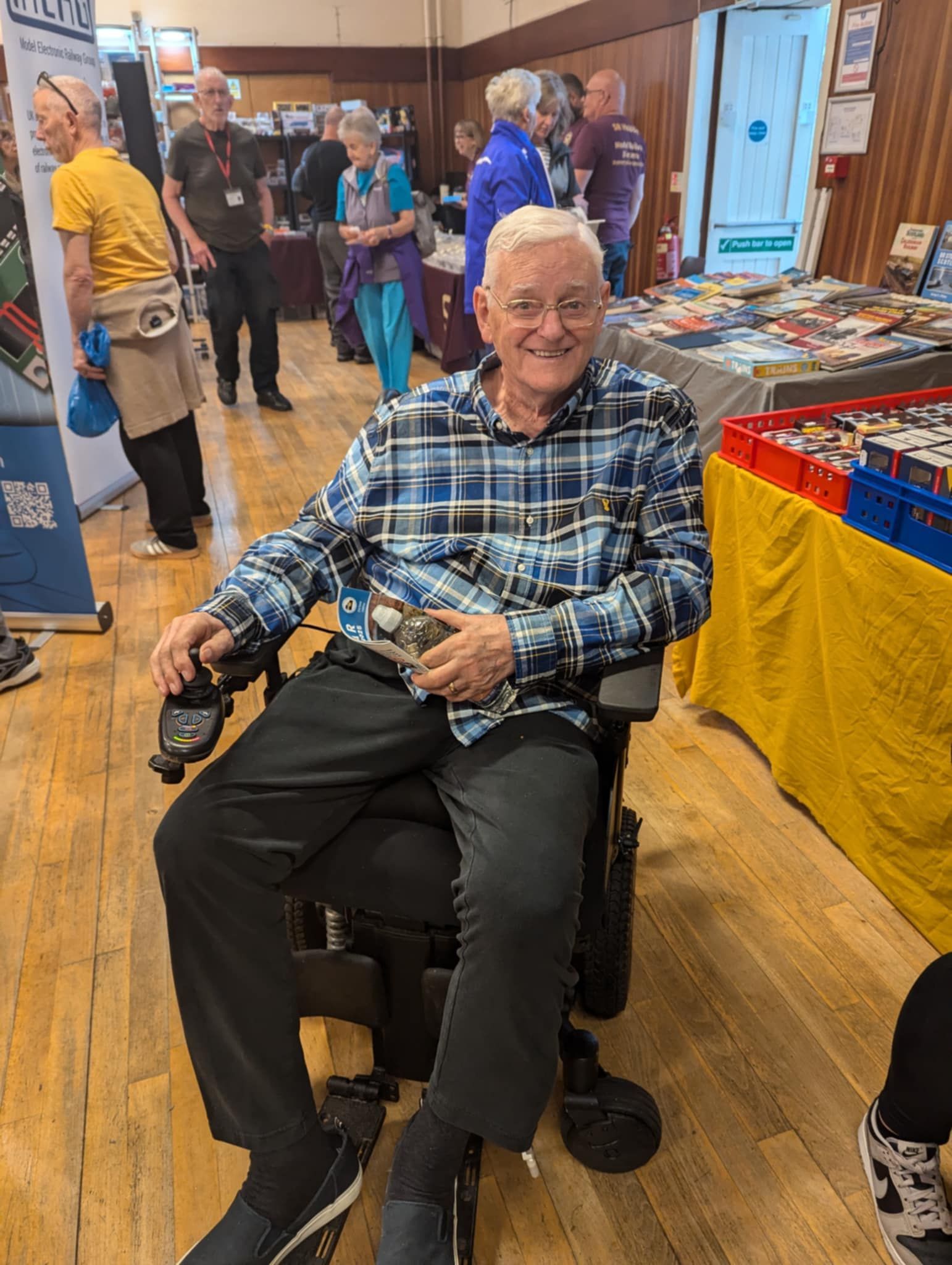 Elderly man in a wheelchair smiling, wearing blue plaid shirt, at an indoor event with vendor tables.