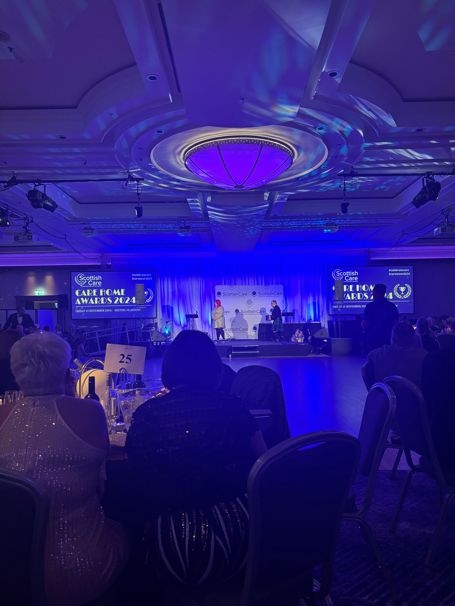 A gala event with a stage, screens, and tables. Blue lighting fills the room. People are seated and enjoying the event.