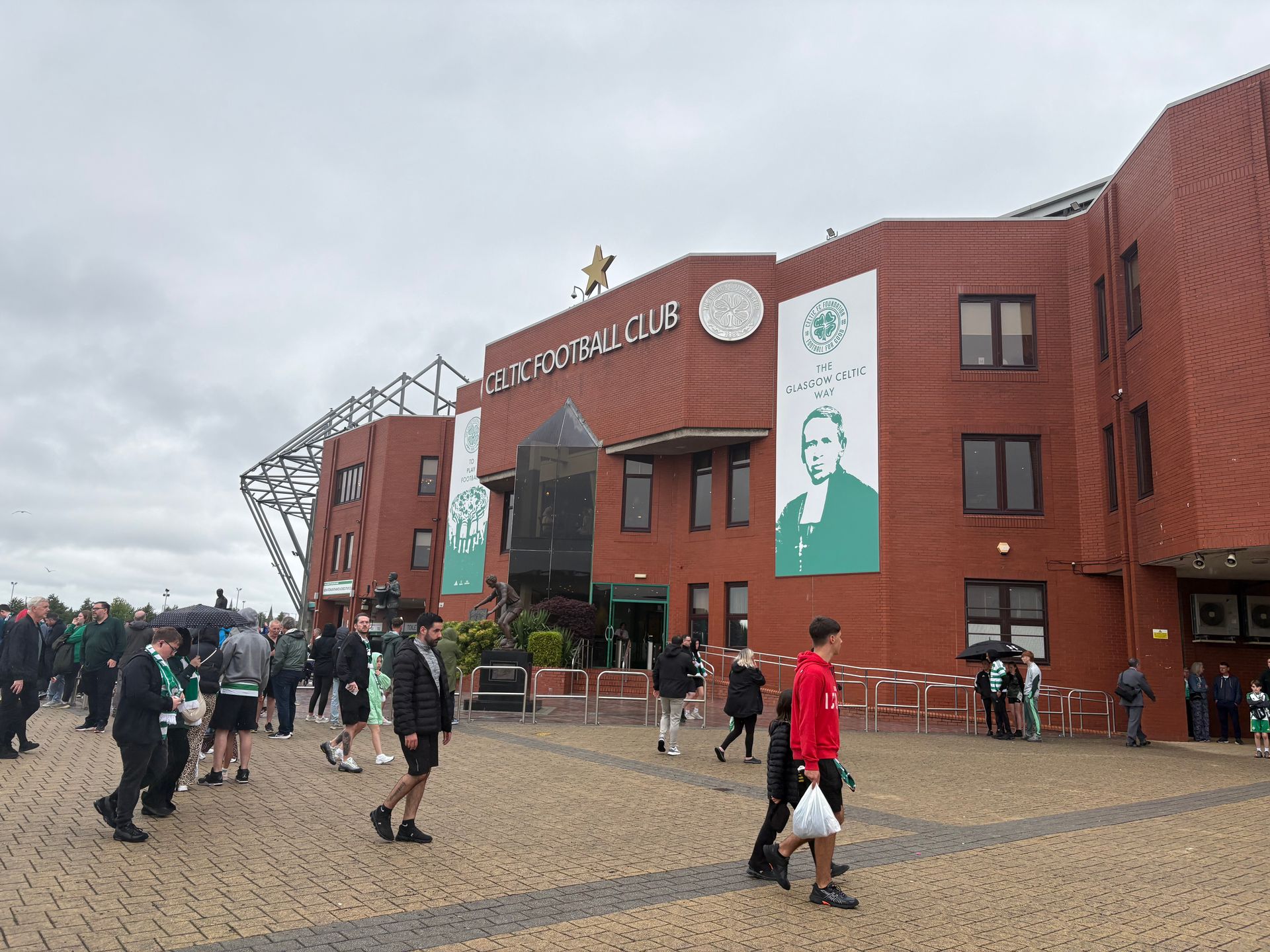 Exterior view of Celtic Park stadium with people in green and white attire milling around.