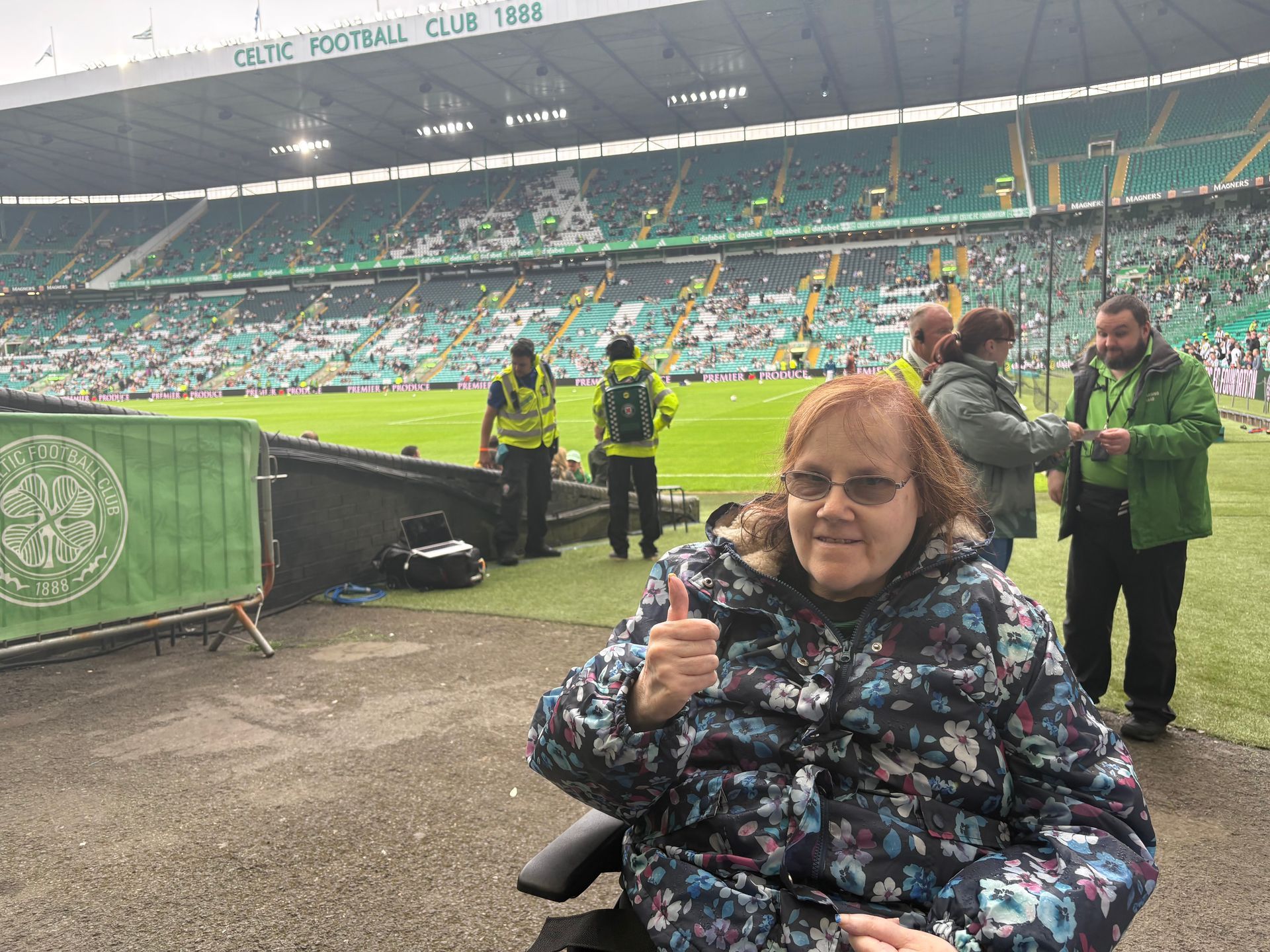 Woman in wheelchair at Celtic FC stadium gives thumbs up. Green and white stadium in background.