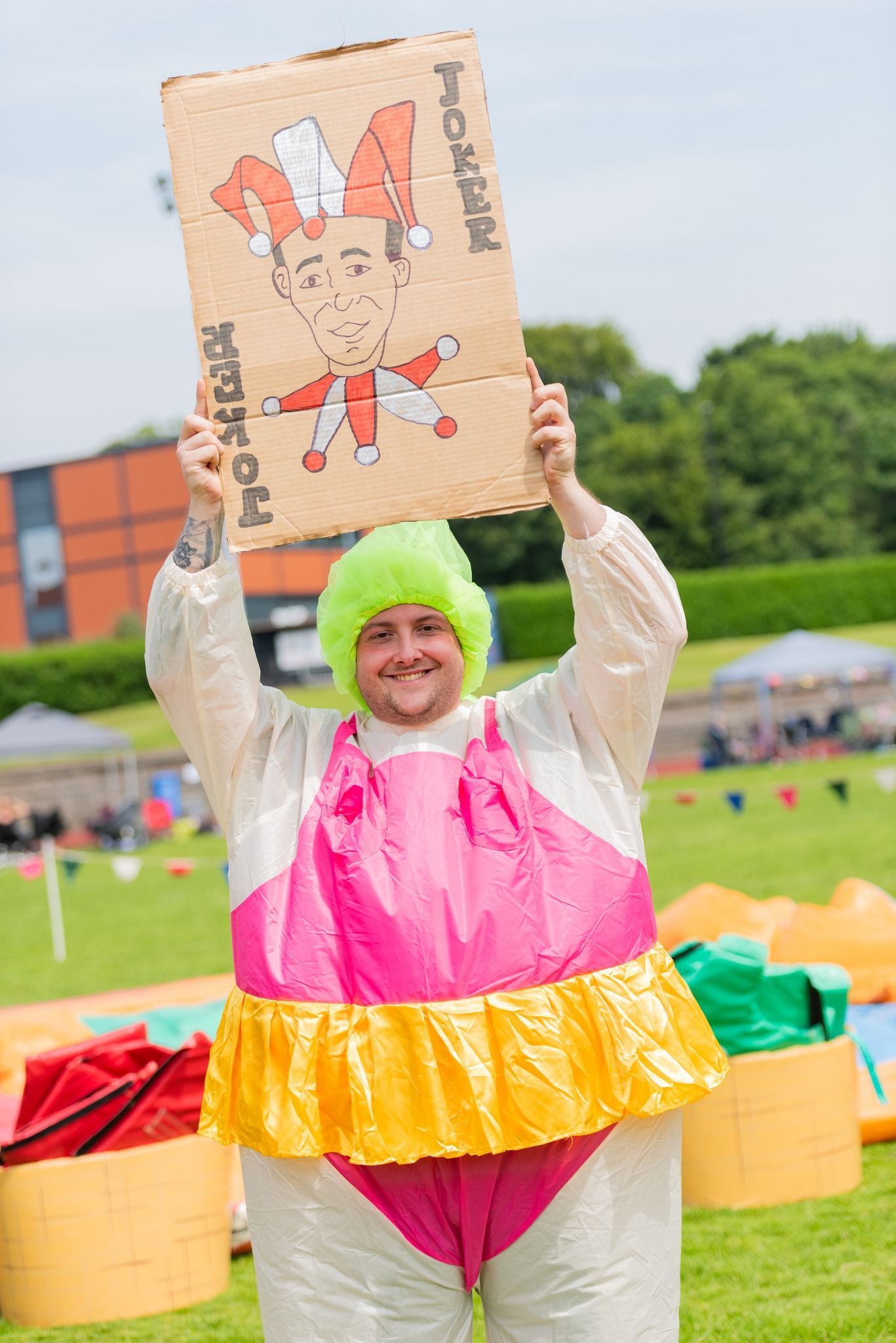 A smiling person in a sumo suit and neon green wig holds up a cardboard sign outdoors. The sign depicts a cartoon character.