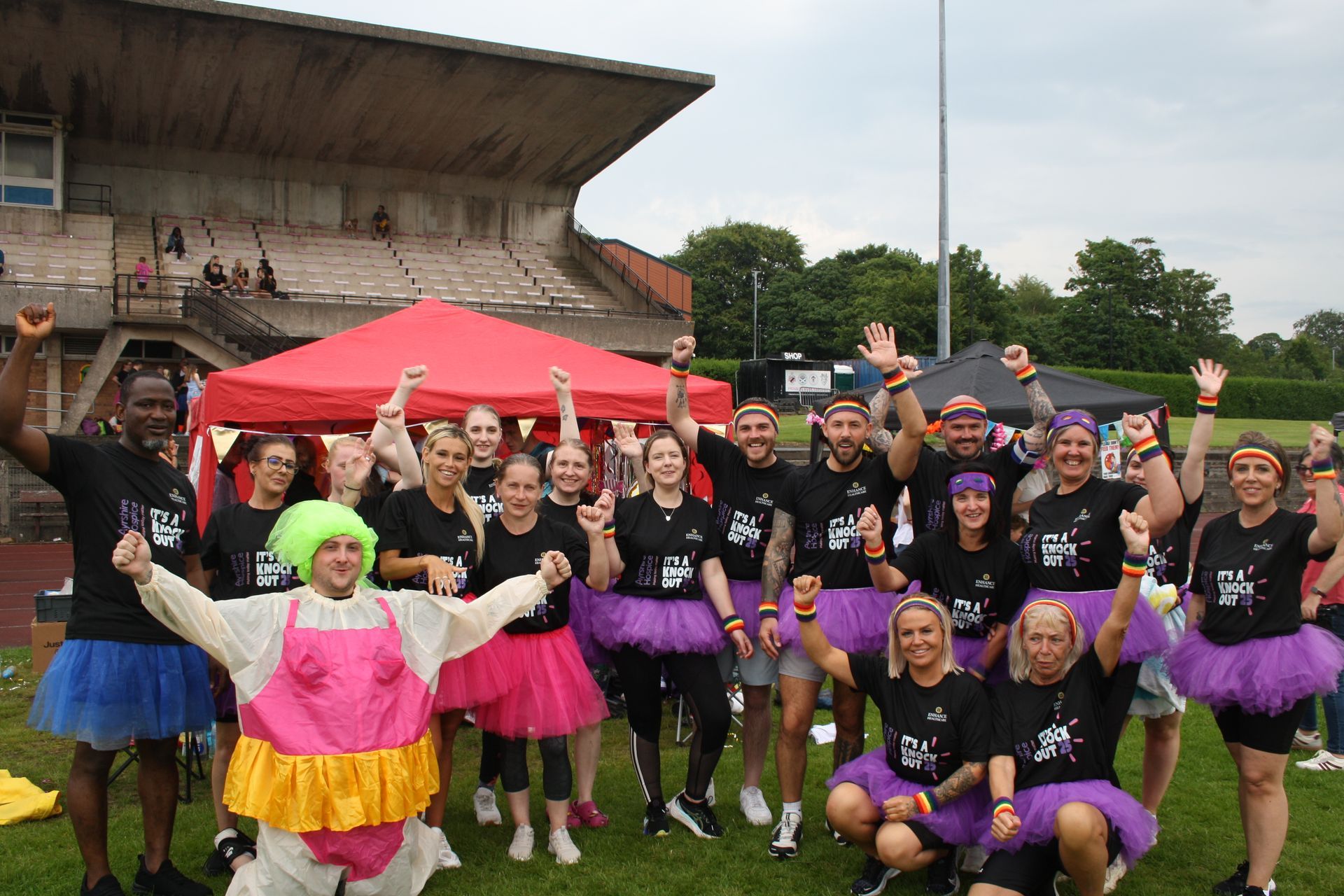Group of people in colorful tutus and black shirts with arms raised, smiling in front of a red tent and stadium seating.