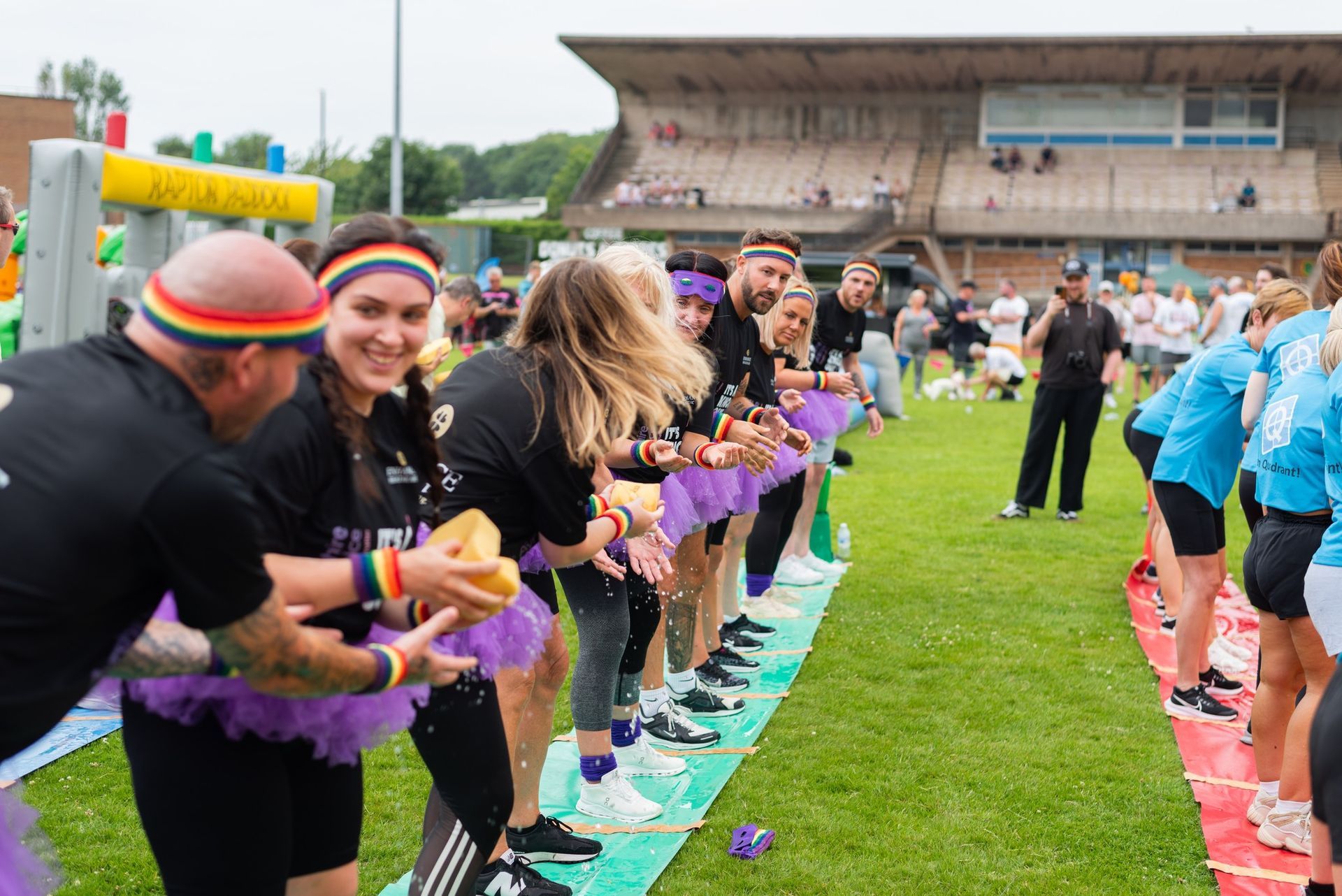 People in purple tutus and athletic wear participate in an outdoor water game, laughing and passing water balloons on a green field.