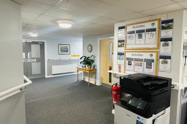 Hallway with information boards, printer, and doors. Carpeted floor, white walls, and a small table with a plant.