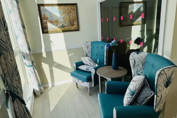 Two teal armchairs with floral patterned cushions and curtains next to a small round table in a sunlit room.